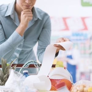 woman with her hand on her chin, standing in front of a shopping cart and looking at a long grocery list