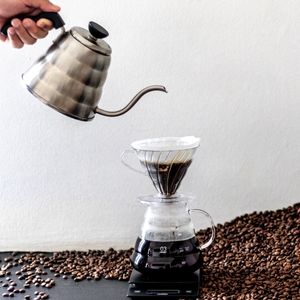 a hand pouring water over a pour-over coffee system surrounded by whole coffee beans